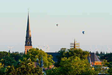 Fototapeta premium European cathedral tower surrounded by green trees at sunset with hot air balloons. Breda, the netherlands