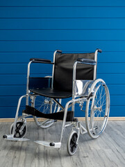 Empty steel wheelchair with black leather seat and backrest on the blue wooden plank wall background, vertical style. Chair with wheels for use as transport by person who unable walk, illness, injury.