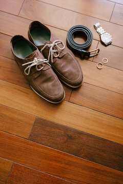 Top view of brown men's shoes with a wristwatch, belt and wedding rings of the bride and groom on a wooden texture.
