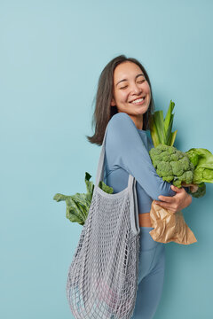 Vertical shot of cheerful brunette Aisan woman stands sideways carries net bag with green vegetables buys products for preparing vegetarian meal smiles happily isolated over blue background.