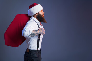 Young bearded man in Santa's hat carries big sack with presents looking ahead. Christmas sales.