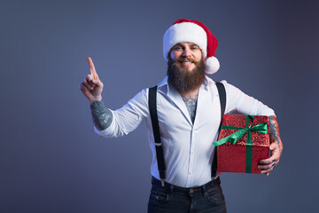 Fototapeta na wymiar Handsome bearded man in Christmas hat holds gift box, shows by index finger up. New Year concept