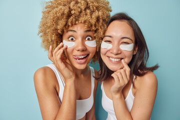 Fototapeta na wymiar Pretty two female friends use undereye skin beauty masks prevent bags reduce facial puffiness enjoy daily skincare routine dressed casually stand indoor against blue background. Beauty procedures