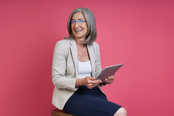 Happy senior woman in formalwear looking away while using digital tablet against pink background