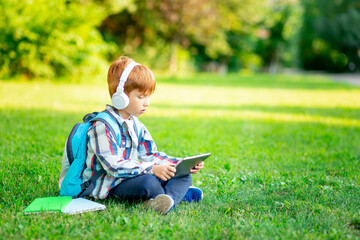 a first-grader boy with a backpack and a tablet with headphones on a green lawn reading a book or...