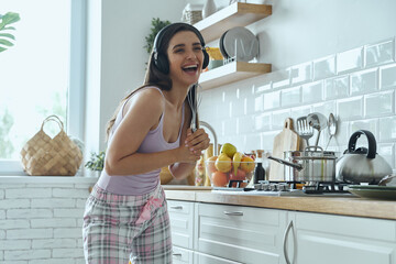 Playful young woman using whisk as microphone and smiling while cooking at the kitchen