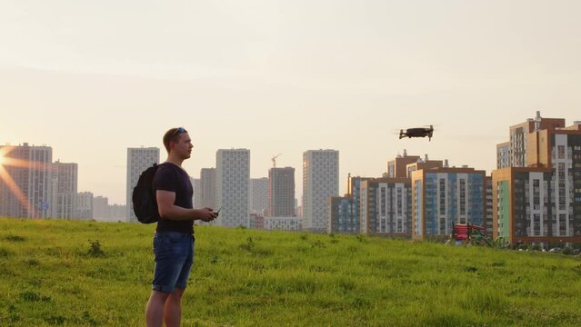 Guy with backpack operates with remote control aerial mobile drone standing at lawn in residential area. Young man managing smart quadcopter for video, photo shooting in summer at cityscape