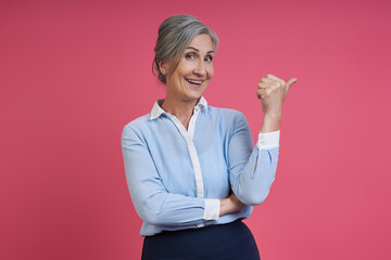 Happy senior woman pointing away while standing against pink background