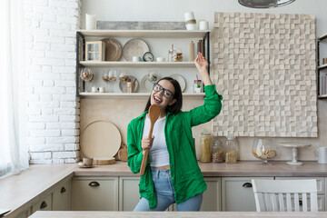 Young beautiful woman in glasses and green shirt in the kitchen having fun relaxing celebrating dancing and singing, brunette happy at everyday work