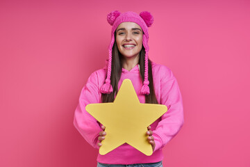 Happy young woman in funky hat holding paper star and smiling against pink background