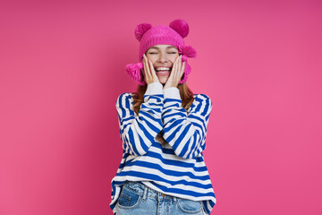 Playful young woman in pink hat touching face and smiling while standing against colored background