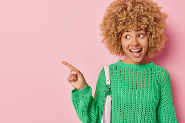 Studio shot of surprised excited young woman with curly hair dressed in casual green jumper carries bag points away on blank space for your advertising content isolated over pink background.