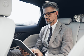 Confident mature businessman working on digital tablet while sitting on the back seat of a car