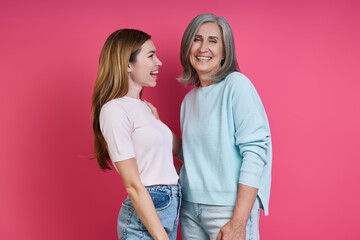 Happy mother and adult daughter standing together against pink background