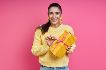 Happy young woman opening gift box while standing against colored background