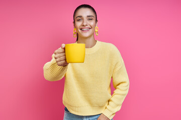 Attractive young woman in yellow sweater holding coffee cup and smiling against colored background