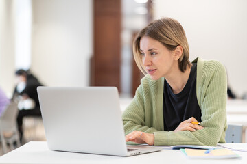 Smiling middle-aged woman studying online on laptop computer while sitting in classroom, doing second degree as mature student. Happy 45s female attending online professional development course
