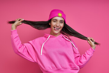 Playful young woman in hooded shirt holding her long hair against pink background