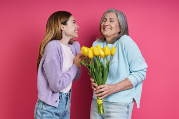 Happy mother holding tulips while her adult daughter standing near and against pink background