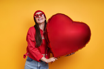 Beautiful young woman holding heart shape balloon and smiling against yellow background