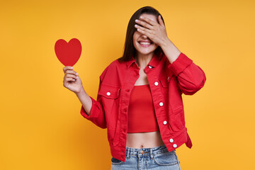 Beautiful young woman holding paper heart and covering eyes with hand against yellow background