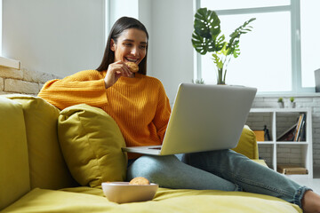 Attractive young woman using laptop and enjoying cookies while sitting on the couch at home