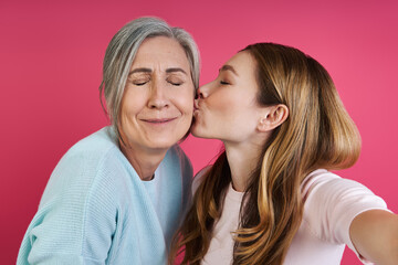Adult daughter kissing her senior mother while making selfie against pink background