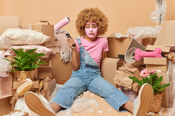 Tired woman with curly hair holds paint roller for doing repair applies beauty mask on face dressed in casual clothes poses on floor in new apartment surrounded by cardboard boxes. Renting concept