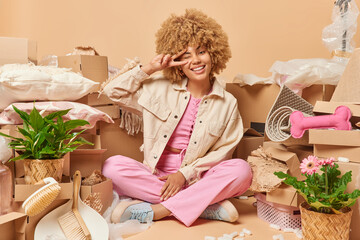 Happy young curly haired woman sits in lotus pose on floor makes peace gesture over eye enjoys starting new life in own apartment relocates and collects belongings in carton boxes. Moving concept