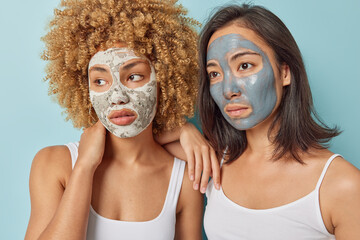 Indoor shot of mixed race young women apply beauty masks for skin treatment focused away with thoughtful expressions dressed in white t shirts isolated over blue background. Wellness concept