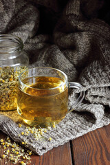 Obraz na płótnie Canvas Camomile herbal dried tea in a glass cup with flowers and knitted woollen sweater on wooden rustic background, closeup, winter cold healing drink, natural medicine and naturopathy concept