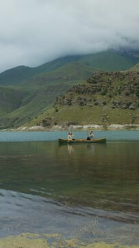 A young couple are sailing in rowboat, across lake against backdrop of mountains