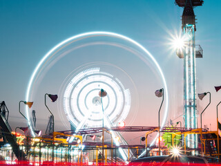 Illuminated ferris wheel, carousels and rides in night. Summer fun for the whole family in the amusement park. Night shot with long exposure and movement of light