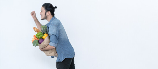 Panoramic banner image of young Asian bearded man hold paper shopping bag full of fresh vegetables and jumping running isolated on white background. Carrying a healthy bag. Web page with copy space. - Powered by Adobe