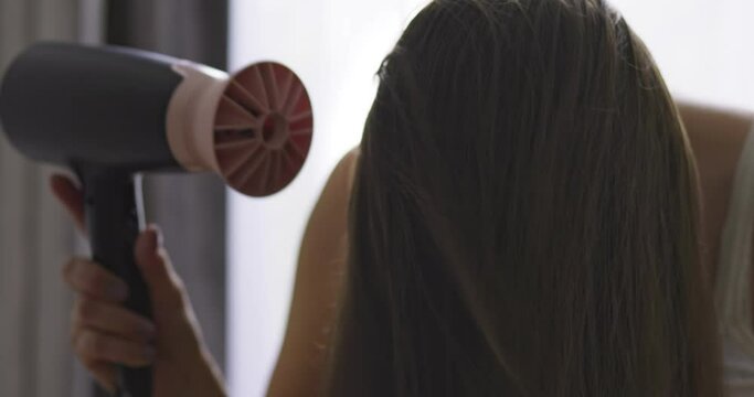 Beautiful young woman using hair dryer near the window at home