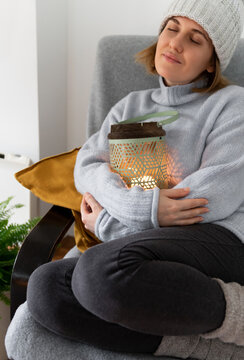 A woman in warm clothes sits in an armchair and hugs a lantern with candle. Low temperature in the house. Energy crisis concept