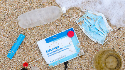 Used reusable face mask with plastic, antigen rapid test kit and other trash lies on sandy beach. Garbage on the ocean shore is washed away by sea foam. Top view