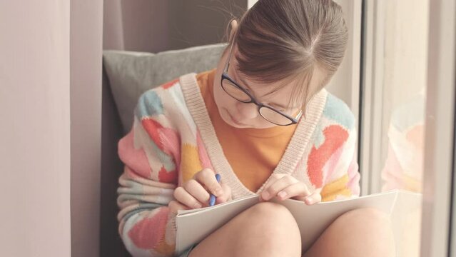 Talented little girl with down syndrome drawing with wax crayons while sitting on windowsill at home