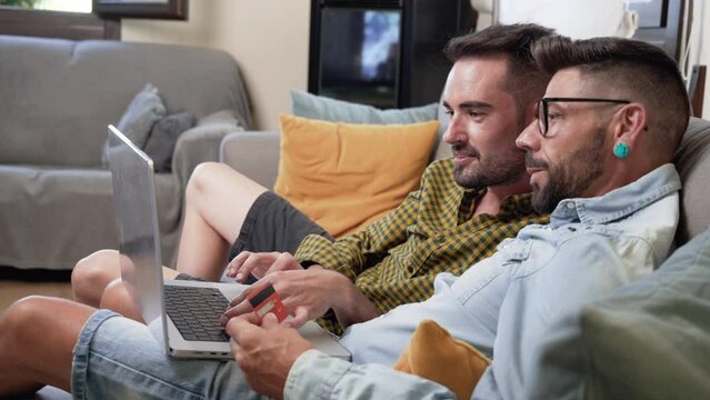 Happy young gay couple using laptop computer while sitting on a couch at home, shopping online with a credit card. High quality 4k footage