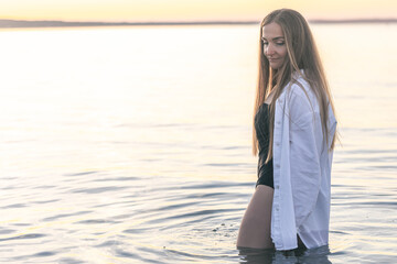 A woman in a swimsuit and a white shirt in the sea at sunset.