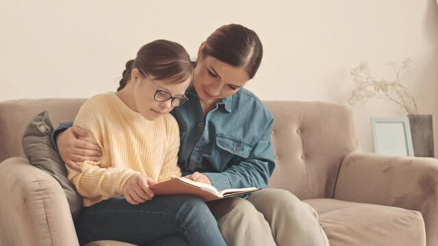 Loving mother and her little daughter with down syndrome sitting on sofa in living room reading book