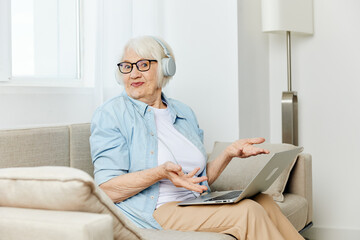 a happy modern old lady looks at the monitor holding a laptop on her lap talking through headphones smiling happily and gesticulating with her hands