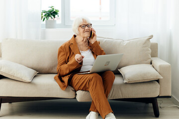 a sad, angry elderly woman in a brown suit is sitting with a laptop on her lap on a cozy sofa working from home and talking on the phone, looking serious and holding her hand in her fist - Powered by Adobe