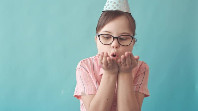Slowmo of cheerful 12 year old Caucasian girl with down syndrome blowing confetti at camera standing on light blue background wearing birthday hat