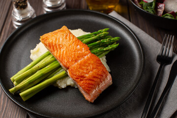 Fried salmon steak is served on a black flat plate, which lies on a pillow of mashed potatoes with parmesan and freshly boiled asparagus, Black cutlery.