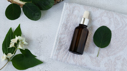 a bottle of dark glossy glass with a pipette lies on a light background on a towel surrounded by green leaves and jasmine flowers. close-up, blank for designers, layout, mock-up