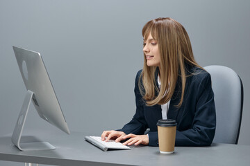 Millennial blonde businesswoman worker in blue jacket using desktop computer with takeaway coffee sitting at workplace in gray office. Remote Job, Technology And Career Profession Concept. Copy space