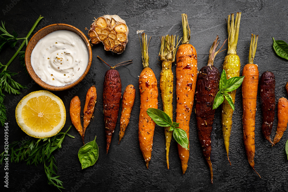 Poster roasted young whole colorful carrot with herbs over black background. top view