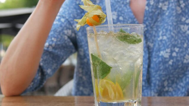 Summer cocktail. Mojito cocktail in a transparent glass glass with pieces of ice, a mint and lemon leaf, a woman drinks a cooling cocktail from a cocktail tube on the veranda of a summer cafe