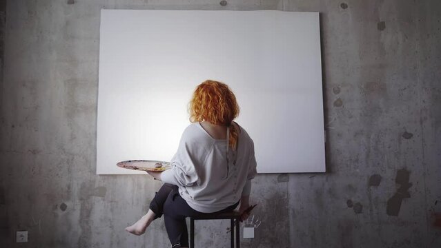 Caucasian artist waiting for inspiration concept. Redhead female painter sitting in front of white canvas holding paintbrushes and palette. High quality 4k footage